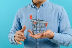 Young smiling man holding a mini shopping cart on a blue background. Sales concept. Place for text. photo