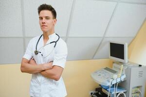 Charming young male doctor , sitting at his office near ultrasound scanning machine. Handsome friendly doctor enjoying working at the hospital, copy space. Ultrasonic concept photo