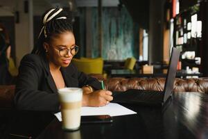 estudiante niña hojeada Internet, utilizando gratis Wifi a cafetería. africano persona de libre dedicación pensando en ideas para su Blog, utilizando ordenador portátil a trabajo colaborativo espacio, descansando mano en de madera mesa, mirando con inspirado sonrisa foto