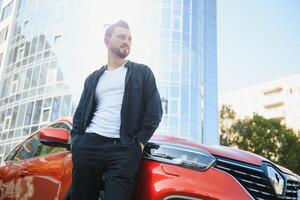 un atractivo hombre con un barba soportes siguiente a su coche en el estacionamiento lote. él es considerado, y mira lejos, alguien es espera. verano día foto