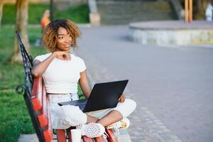 Beautiful smiling young african woman sitting on a bench outdoors, working on laptop computer photo