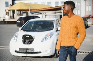 Urban, electric vehicle, eco concept. Young black skinned man, waiting for his electric car charging photo