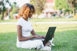 Thoughtful cute mixed female international student with curly hair is sitting on fresh grass with modern laptop in public park, leaning on apple tree and wistfully looking aside during her break photo