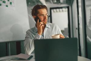 empresario hablando teléfono con cliente mientras sentado en trabajo colaborativo con ordenador portátil y Bebiendo café foto