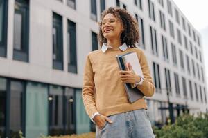 Smiling female freelancer standing with laptop and note pads on modern building backgrounds photo