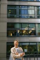 Business woman standing with laptop and coffee on office terrace during break time and looks away photo