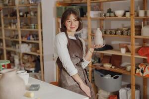 Portrait of young female potter in apron with mug looks away while posing in workshop photo