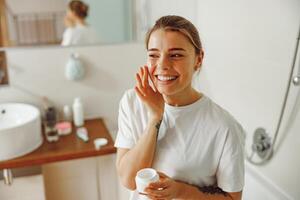 sonriente joven mujer tomando cuidado de piel por aplicando hidratante crema en baño foto