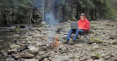 A man works on a laptop near a stream in nature. Concept of freelancing, digital nomad or remote office. 4K video