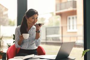 Woman manager recording audio message on mobile phone during coffee break in office photo