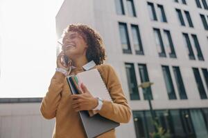 Smiling woman manager is talking phone while standing with laptop on modern building background photo
