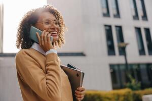 Smiling business woman is talking phone while standing with laptop on modern building background photo
