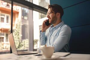 masculino jefe hablando teléfono con cliente mientras sentado en moderno oficina y trabajando en ordenador portátil foto