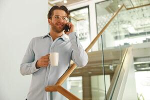 masculino ventas gerente con café taza hablando teléfono con cliente mientras en pie en trabajo colaborativo en escalera foto