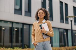 Young smiling female student standing with laptop and note pads on modern building backgrounds photo