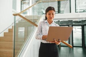 Smiling woman sales manager working on laptop while standing on stairs in coworking background photo