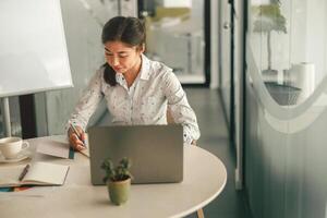 sonriente mujer oficina gerente haciendo notas mientras trabajando en ordenador portátil en moderno trabajo colaborativo foto
