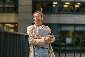 Business woman standing with laptop and coffee on office terrace during break time and looks away photo