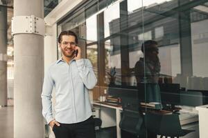 Handsome male entrepreneur talking phone with client while standing in office and looking at camera photo