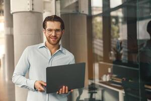 elegante empresario con ordenador portátil en pie en moderno oficina antecedentes y mirando a cámara foto