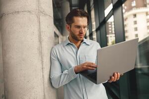 Focused male sales manager in eyeglasses working on laptop standing in office during working day photo
