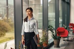 Portrait of stylish business woman standing with laptop near office window and looking at camera photo