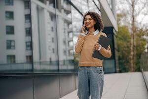 Pretty female entrepreneur is talking phone while standing with laptop on modern building background photo