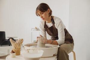 Concentrated female artisan in apron sitting on bench with pottery wheel and making clay pot photo