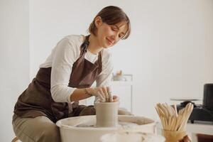 Smiling female artisan in apron sitting on bench with pottery wheel and making clay pot photo