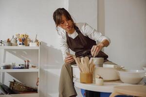 Focused female artisan in apron sitting on bench with pottery wheel and making clay pot photo