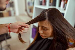 Young female customer sits in a chair covered with a cape while a hair master cuts fringe photo