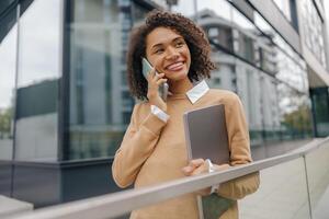 Smiling woman manager is talking phone while standing with laptop on modern building background photo