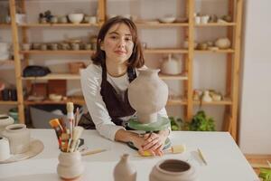 Portrait of young female potter in apron with mug looking at camera while posing in workshop photo