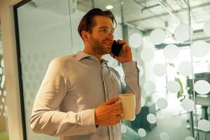 Male sales manager with coffee cup talking phone with client while standing in coworking background photo