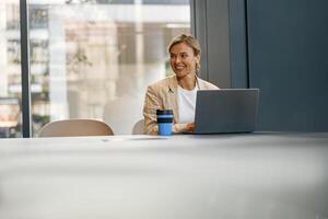 Smiling female entrepreneur working on laptop sitting the desk on office background and looks away photo