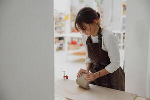 Professional female potter in apron kneads piece of clay with her hands on table in studio photo