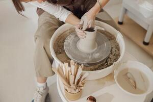 Female potter in apron making shape of clay vase on spinning pottery tool in ceramic workshop photo