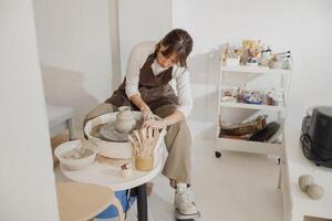 Focused female artisan in apron sitting on bench with pottery wheel and making clay pot photo