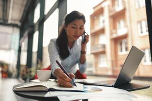 Woman office manager talking phone with client and making notes while sitting the desk in office photo