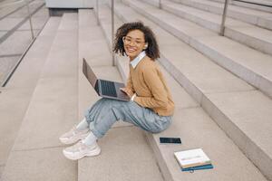 sonriente joven hembra estudiante trabajando en ordenador portátil sentado en escalera en moderno edificio antecedentes foto