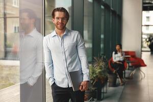 Smiling manager with laptop standing in office hall on colleague background and looking at camera photo