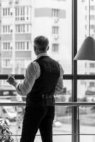 Man staying near big window. Male in suit looking ahead. View from the back. Panoramic city view. Relaxing and taking break. Black and white photo. photo