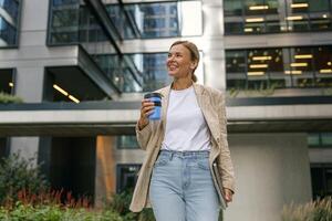 Business woman with laptop drinking coffee during break time near office building and looks away photo