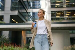 negocio mujer con ordenador portátil Bebiendo café durante descanso hora cerca oficina edificio y mira lejos foto