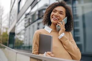 Smiling woman manager is talking phone while standing with laptop on modern building background photo