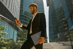 Handsome businessman using mobile phone standing with laptop on background of skyscrapers photo