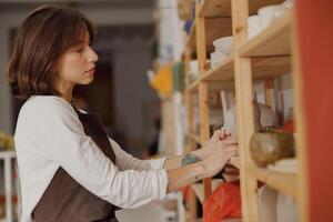 Pretty female ceramist standing near shelves of clay mugs while working in pottery studio photo