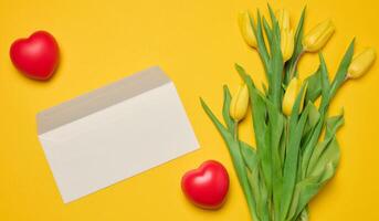 sobre y rojo corazón y ramo de flores de floreciente tulipanes con verde hojas en un amarillo antecedentes foto