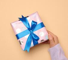 A woman's hand holds a gift box wrapped in a blue silk ribbon on a beige background photo