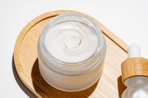 close up of collagen cream in open glass jar and tip of dropper bottle on bamboo tray on white background. Set for skin and body care beauty products photo